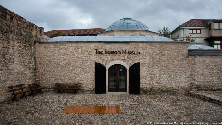 Stone façade of The Hamam Museum with triple-arched entrance, metal-clad dome, benches, and cobbled forecourt.