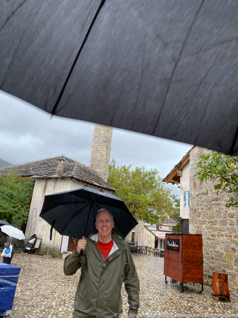 Smiling visitor under a black umbrella in a wet cobbled courtyard with a stone minaret behind.
