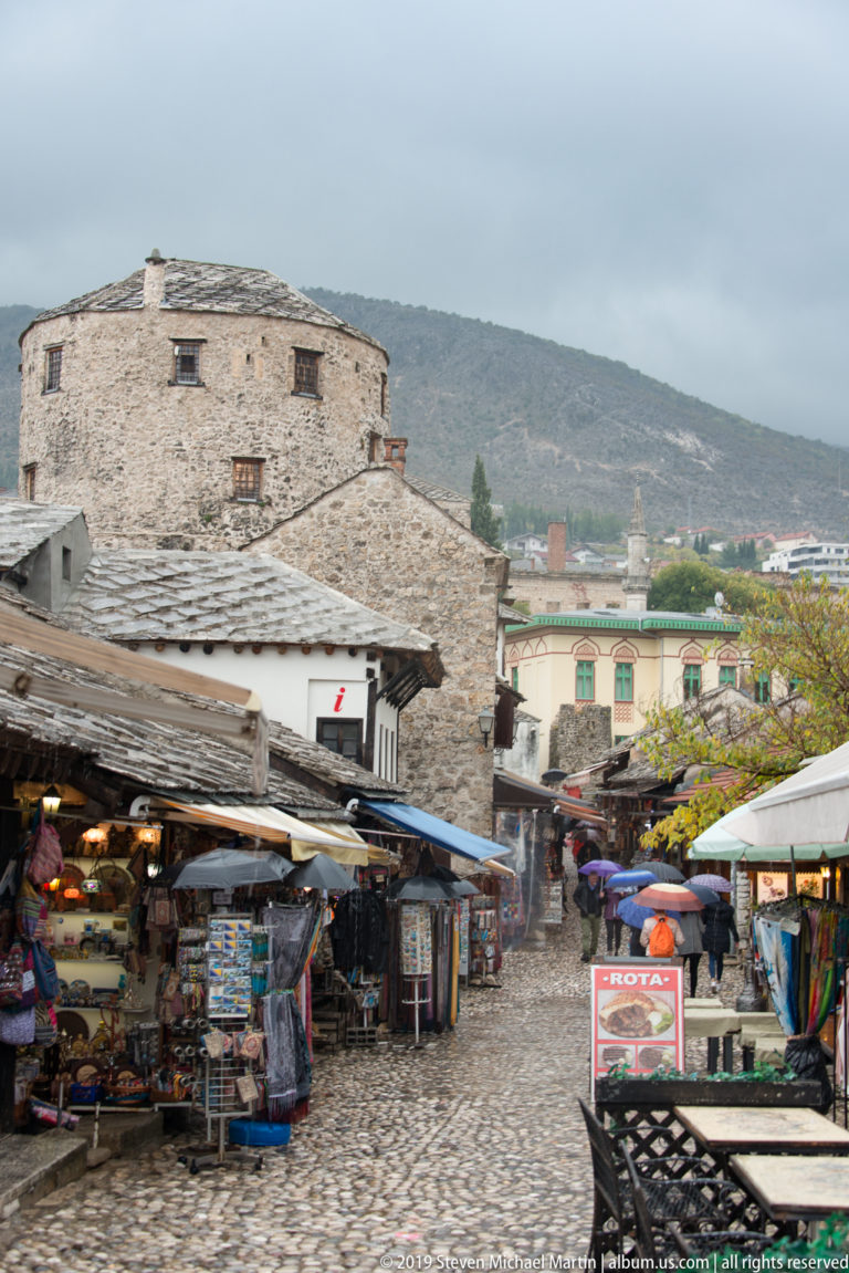 Rainy cobbled lane through Kujundžiluk bazaar with souvenir stalls, stone buildings, awnings, and mountains beyond.