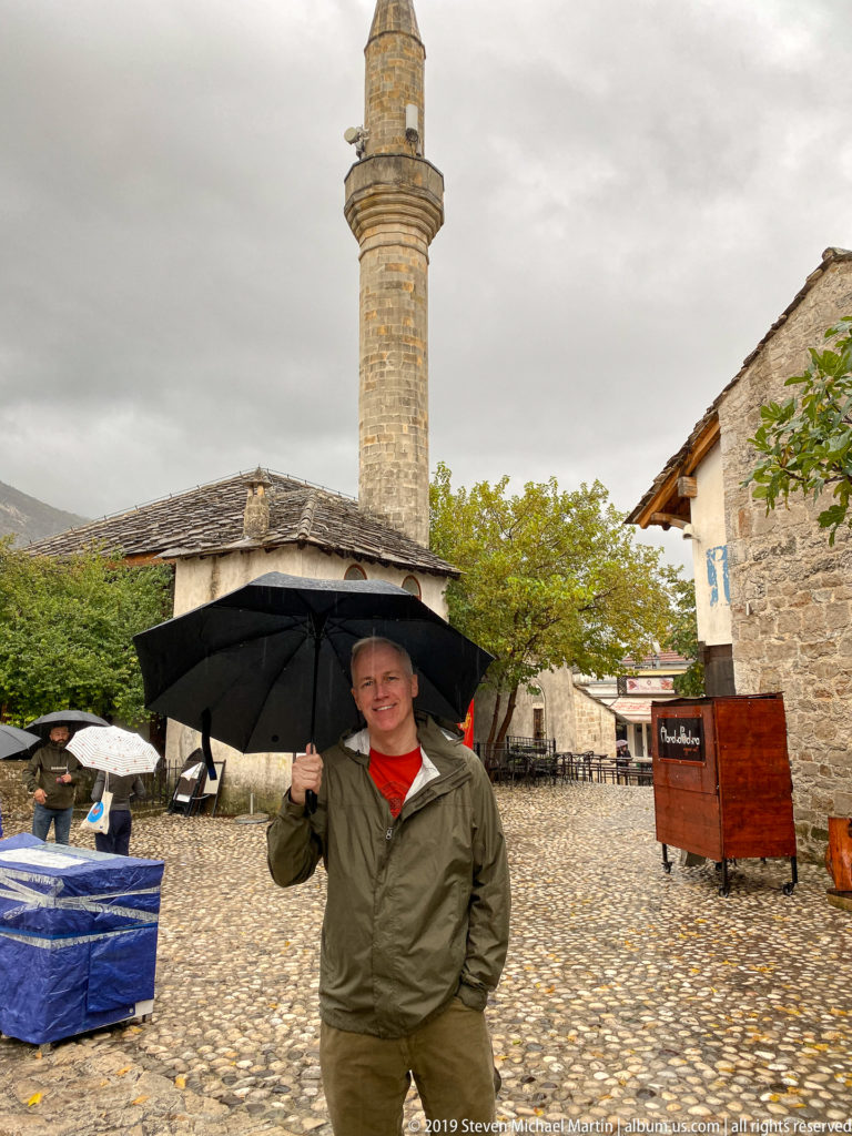 Person holding a black umbrella in a cobbled courtyard beneath a tall stone mosque minaret in Mostar.