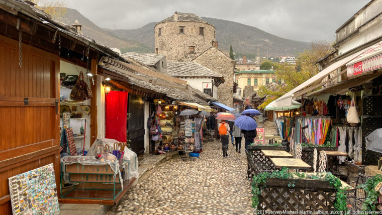 Narrow cobblestone bazaar lane lined with souvenir shops and pedestrians beneath umbrellas in Mostar.