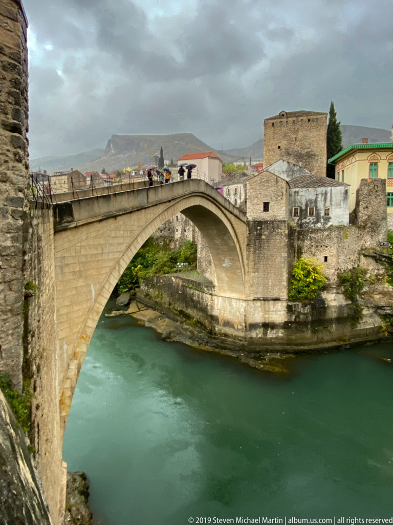 Side view of Stari Most arch over turquoise Neretva water with a stone bridge tower and pedestrians.