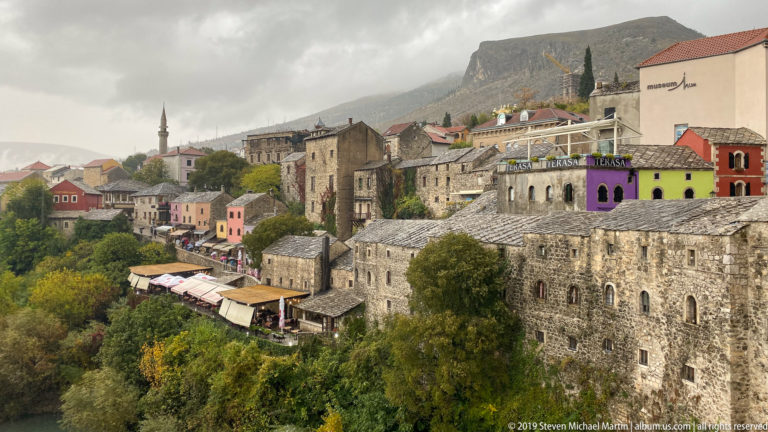 Stone and colorful buildings along Mostar’s wooded riverbank with a minaret and mountain ridge behind.