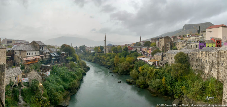 Neretva River running between steep built-up banks, colorful houses, trees, and minarets in Mostar.