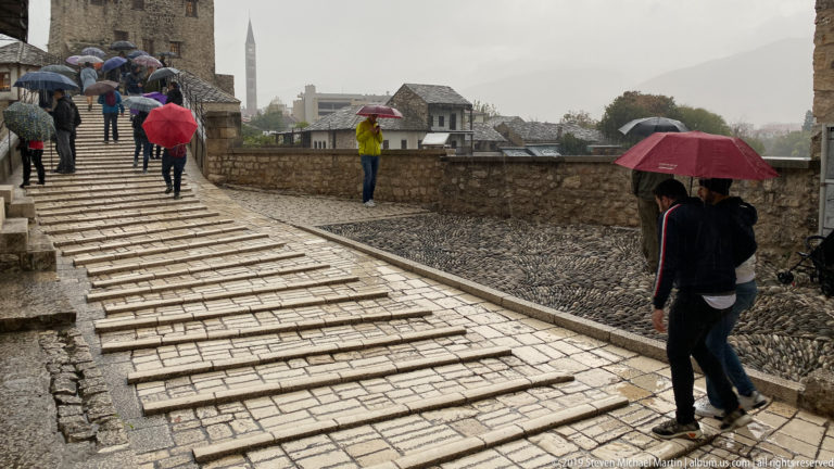 People with umbrellas walking across the wet, steep stone deck of Stari Most.