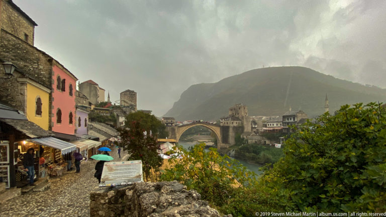 Umbrella-carrying pedestrians on a cobblestone old-town lane with Stari Most visible in the distance.