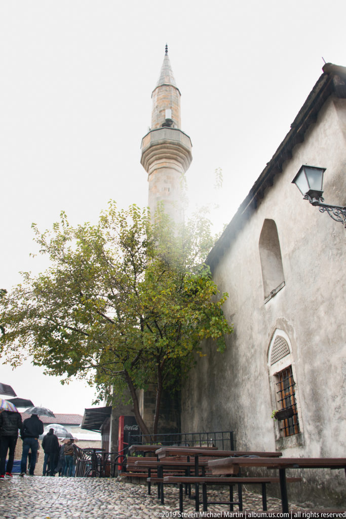 Stone minaret and small plastered mosque beside a leafy courtyard and bazaar pedestrians in Mostar.