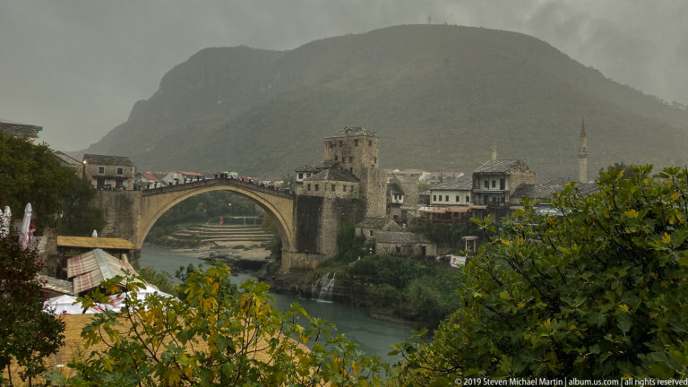 Stari Most stone arch spanning the Neretva River with bridge towers, old-town buildings, and mountain backdrop.