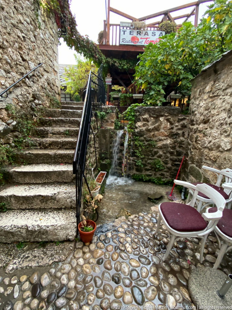 Stone steps and patterned cobblestone courtyard with café chairs and potted plants in Mostar.