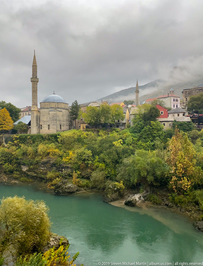 Domed stone mosque and tall minaret above trees beside the Neretva River in Mostar.