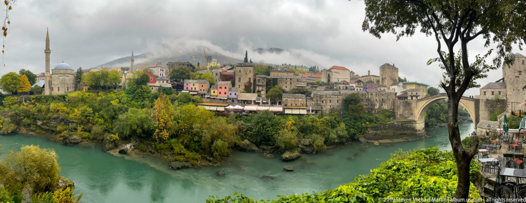 Historic Mostar above the Neretva River with stone buildings, minarets, autumn trees, and bridge-quarter masonry.
