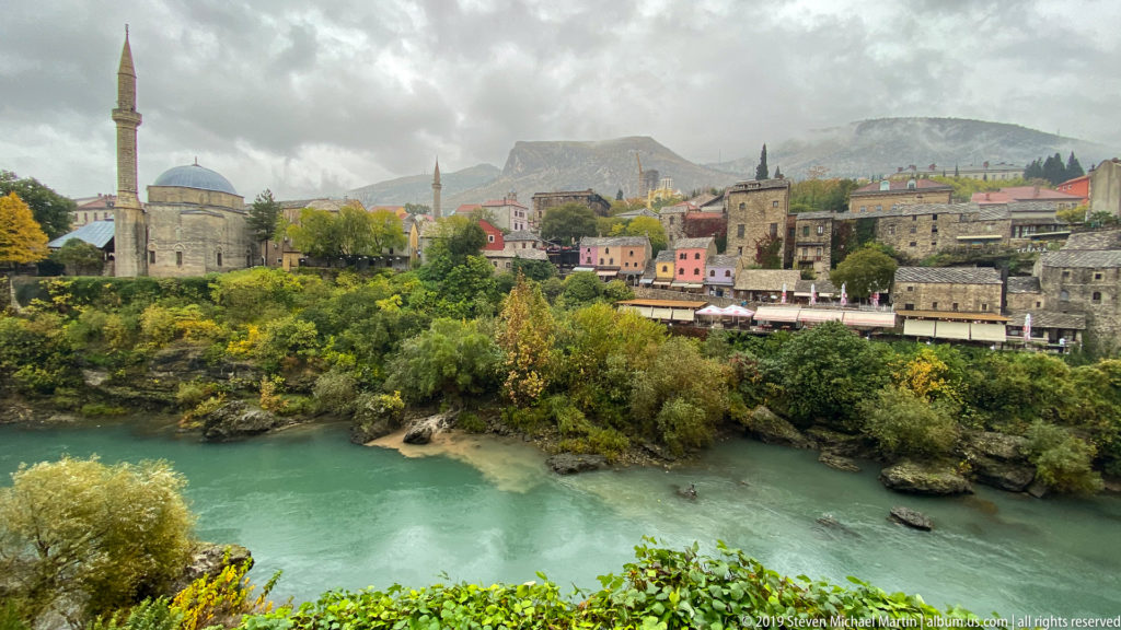 Turquoise Neretva River below historic stone buildings, a domed mosque, minarets, and misty mountains in Mostar.
