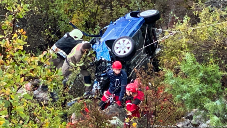 Rescue workers gather below an overturned blue car on a steep wooded slope.