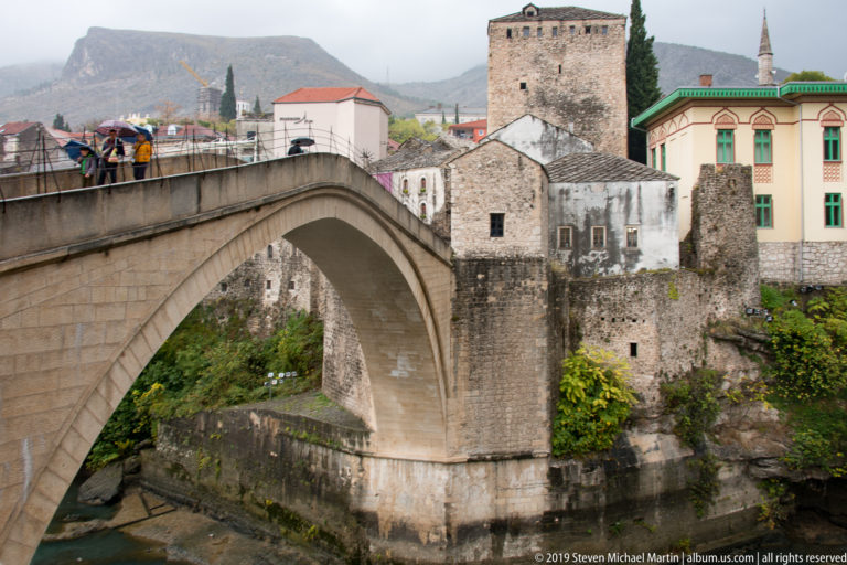 Stari Most arch with pedestrians on the deck, backed by a large square stone tower and historic riverside buildings.