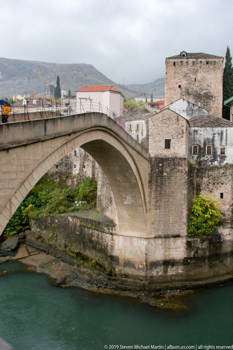 Close side view of Stari Most’s high stone arch above the Neretva, with pedestrians crossing the narrow bridge deck.