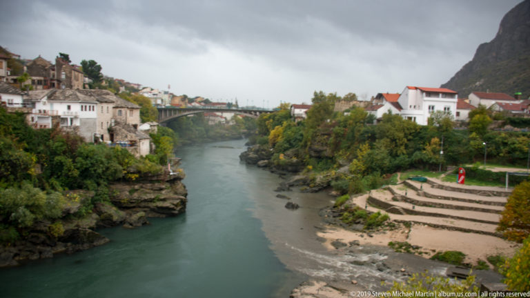 Neretva River flowing toward a distant arch bridge, with stepped terraces, houses, autumn trees, and mountains on both banks.