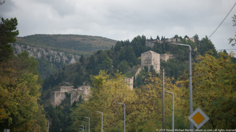 Stone towers of the Vidoški fortress rise through autumn trees on a steep hillside above a road near Stolac.