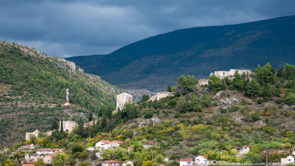 Stone fortification walls and towers descend a rocky hillside above houses, backed by a dark mountain.