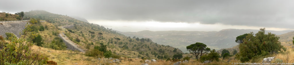 Steep rocky mountain valley with scattered trees and a narrow hillside road beneath low gray clouds.