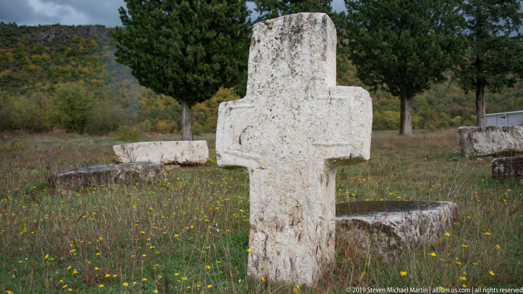 Freestanding pale stone cross stands in flower-dotted grass among low tombstones and trees.