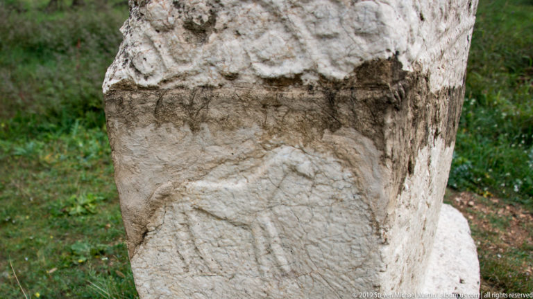 Close view of an eroded human figure carved on a pale stone stećak beneath a projecting edge.