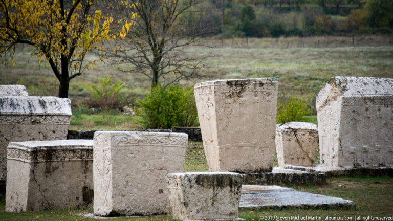 Varied carved stećci stand across a grassy field, including upright, chest-shaped, and gabled forms.