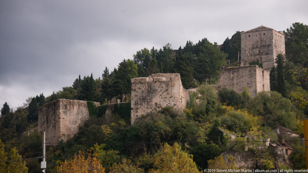 Stone fortress walls and a square tower rise above a wooded hillside under an overcast sky.