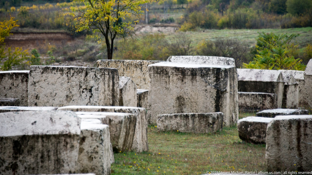 Dense group of rectangular stone stećci across grass, with autumn vegetation and low hills beyond.
