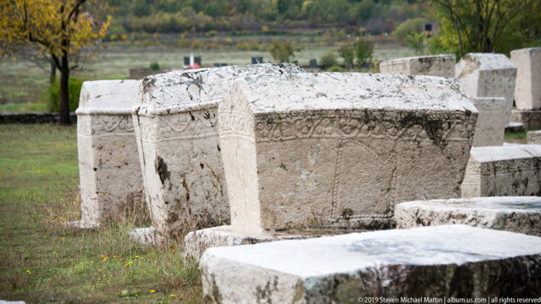 Row of pale carved stećci with gabled tops and ornamental bands recedes diagonally across grass.
