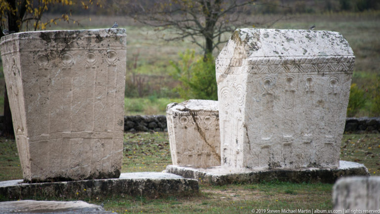 Tall gabled stećak with carved border and circular motif stands among smaller upright stone monuments.