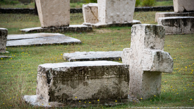 Upright stone cross stands beside a low carved rectangular stećak, with more tombstones in the background.
