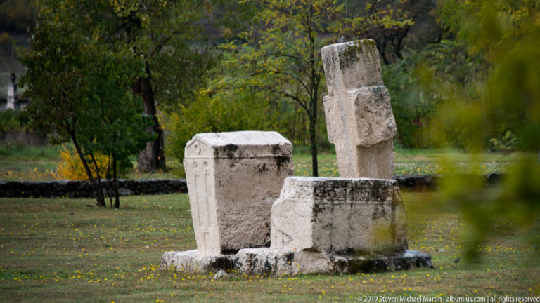 Gabled, chest-shaped, and tall cross-like stone monuments stand together on grass beneath autumn trees.
