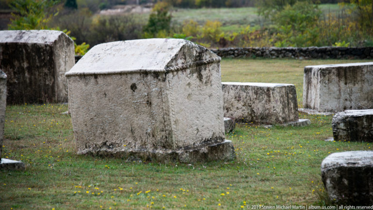 Tall pale stećak with a pitched roof-like top stands among lower stone tombstones on grass.