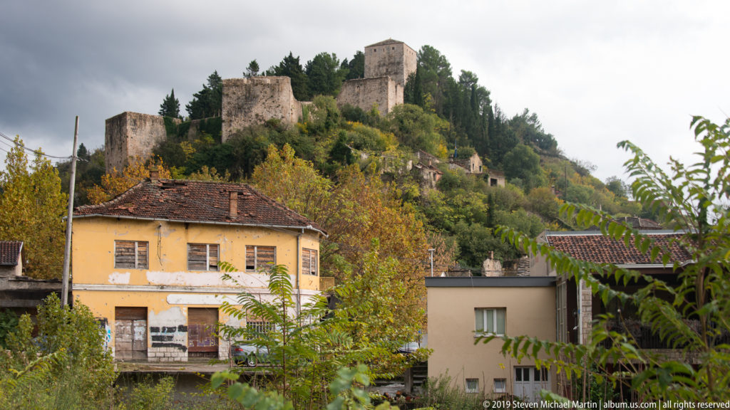 Stone fortress and square tower rise on a wooded hill behind houses with tiled roofs and autumn trees.