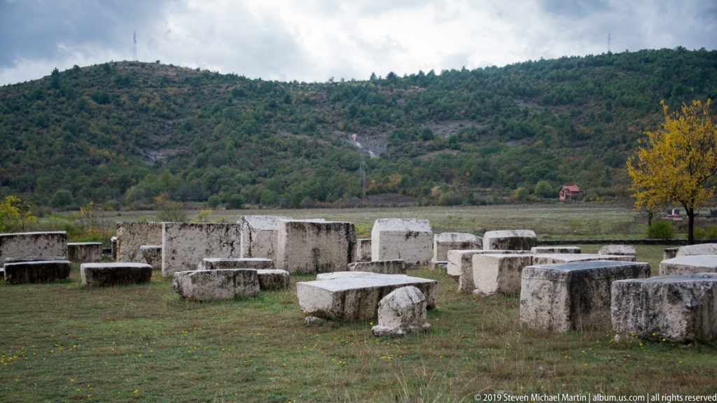 Scattered pale stone stećci fill a grassy necropolis with wooded hills rising in the background.