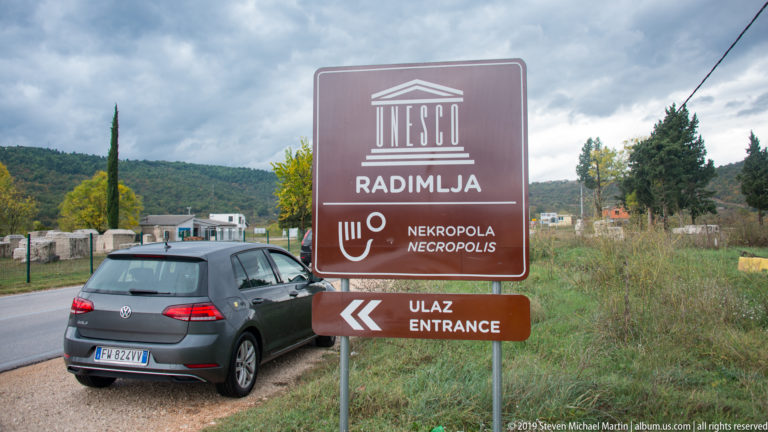 Brown UNESCO Radimlja necropolis entrance sign beside a road, with a parked car and stone monuments beyond.