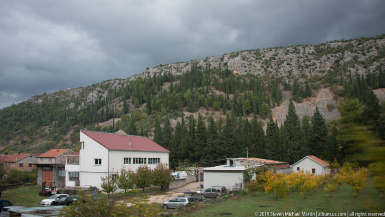 White buildings with red roofs sit below a rocky, tree-covered limestone ridge beneath dark clouds.