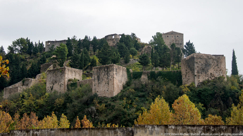 Stone fortress walls and a square tower rise through autumn trees on a steep hill above the town.