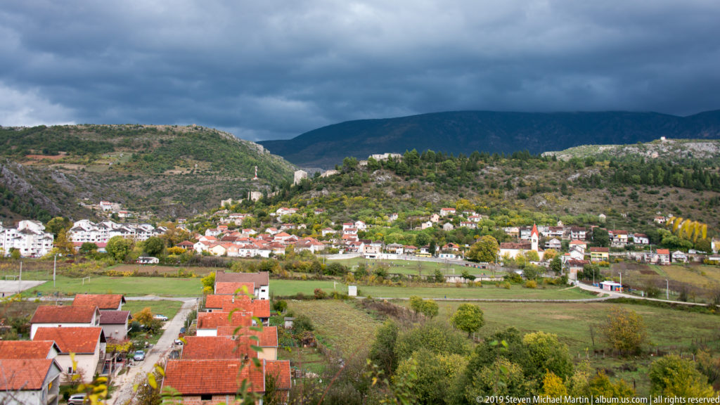 Red-roofed houses and rectangular fields fill a valley below dark mountains and storm clouds.