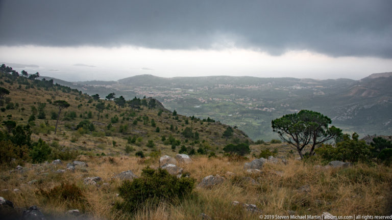 Rocky scrub-covered karst hillside overlooking a hazy valley and distant mountains beneath low clouds.