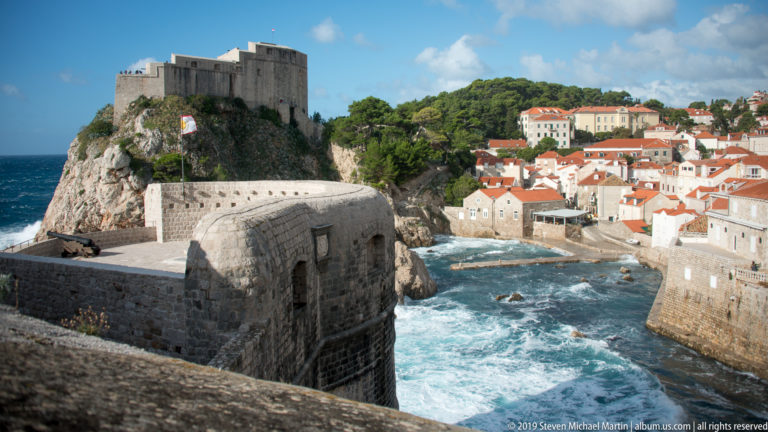 Heavy stone city walls frame a view across rough water to Fort Lovrijenac and red-roofed buildings on the western shore.