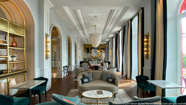 Long upscale hotel lounge with arched wall openings, tall windows, chandeliers, sofas, armchairs, and small marble-topped tables.