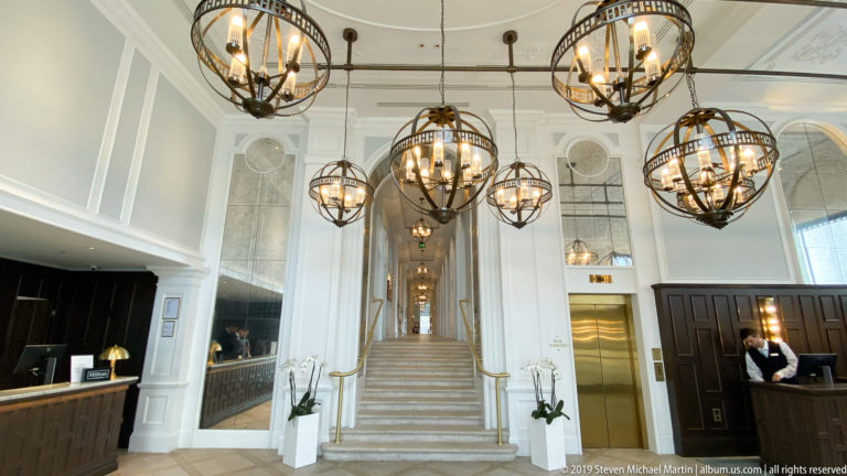 Symmetrical hotel lobby with a broad central staircase, dark wood desks, pale walls, and multiple spherical chandeliers.