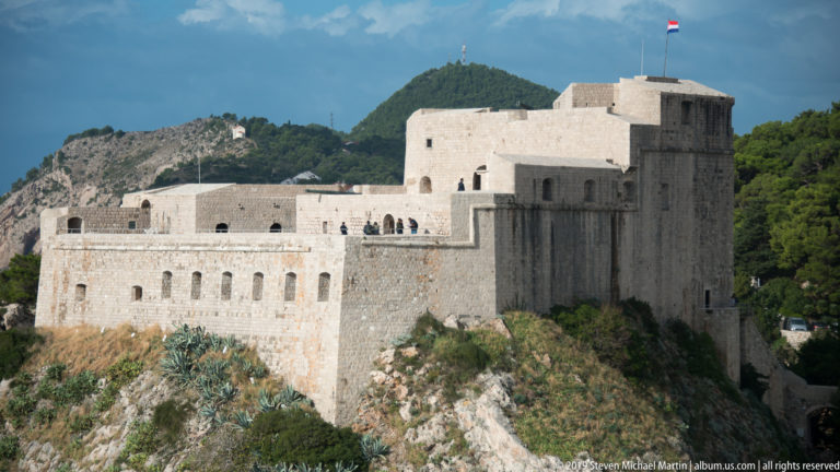 Close view of Fort Lovrijenac’s pale stone walls rising from a rocky cliff, with a Croatian flag flying above.