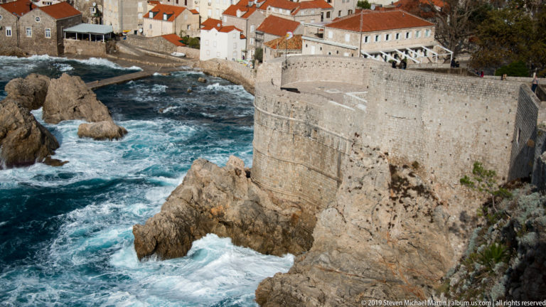 Curved limestone fortification wall towers over jagged rocks and churning blue-white surf along Dubrovnik’s seaward edge.