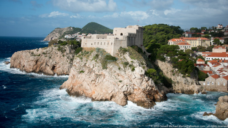 Fort Lovrijenac rises on a steep rocky promontory above dark blue sea, with red-roofed Dubrovnik buildings behind.