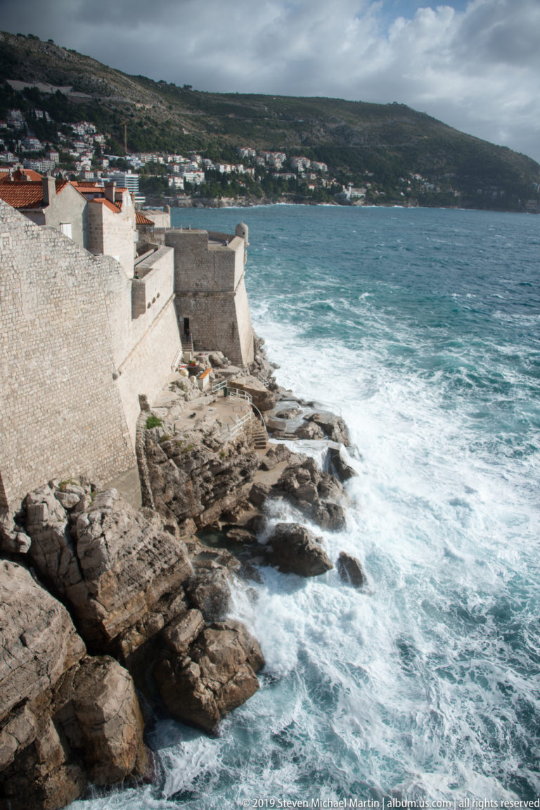 Pale limestone city walls meet jagged coastal rocks as rough Adriatic waves break directly below the fortifications.