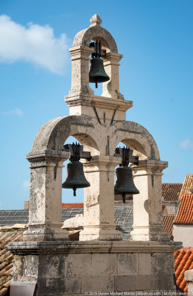 Weathered pale stone bell gable with three large bells and one smaller bell above red-tiled roofs against blue sky.