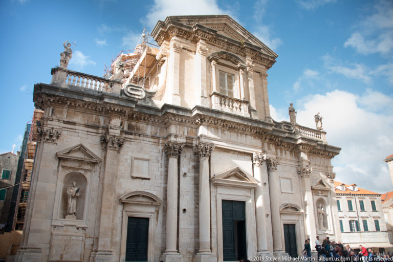 Baroque façade of Dubrovnik Cathedral with pale stone pilasters, pediments, niches, statues, and a raised central bay.