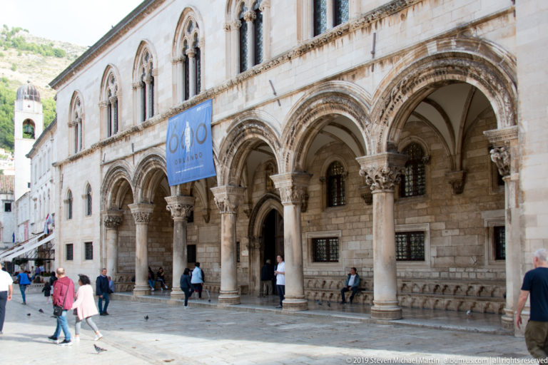Street view of the Rector’s Palace with open stone arcade, carved columns, upper windows, pedestrians, and blue 600 banner.